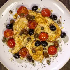 Petto di pollo in padella con pomodorini, olive nere e capperi, condito con erbe aromatiche.