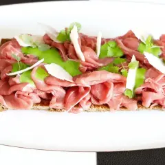 Delicato carpaccio di manzo su crostino integrale, guarnito con crema verde, scaglie di parmigiano e germogli freschi.