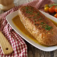 Un succulento polpettone in padella, dorato e profumato, servito in un piatto bianco con erba cipollina e contornato da pomodorini.