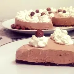 Una fetta di golosa cheesecake alla Nutella, guarnita con panna montata e nocciole intere, su un piatto bianco.