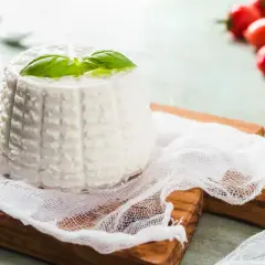 Ricotta fatta in casa, fresca e profumata, guarnita con basilico su un tagliere di legno. Pomodorini rossi sfocati sullo sfondo.