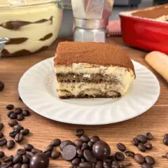 Una fetta di tiramisù con uova pastorizzate, cosparso di cacao, su un piatto bianco, con chicchi di caffè e cioccolatini sparsi.