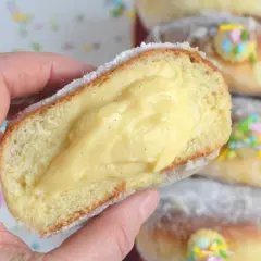 Un bombolone alla crema, soffice e ripieno, viene tenuto in mano. Altri bomboloni alla crema decorati con confettini colorati sono sullo sfondo.