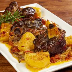 Un succulento agnello al forno con patate dorate, pomodorini e rosmarino su un piatto bianco.