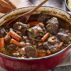 Un invitante spezzatino di manzo in un tegame rosso, con carote, funghi e pane rustico sullo sfondo.
