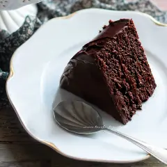Una fetta di torta al cioccolato, umida e golosa, su un piatto bianco con bordo dorato. Accanto, un cucchiaino d'argento.