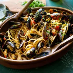 Un invitante piatto di pasta con le cozze, condita con pomodoro e prezzemolo, servita in una ciotola di legno con cucchiai.