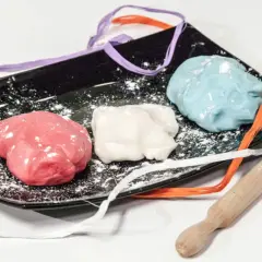 Tre panetti di pasta di zucchero colorata (rosa, bianca, azzurra) su un piatto nero con nastri colorati e un mattarello.