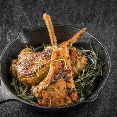 Capretto in padella, dorato e profumato, servito con rosmarino fresco su una piastra di ghisa.