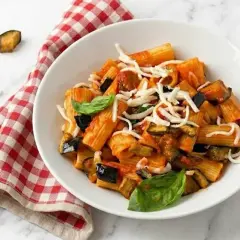 Un piatto di pasta con melanzane, pomodoro e formaggio grattugiato, guarnito con basilico fresco. Accanto, un tovagliolo a quadri rossi e bianchi.