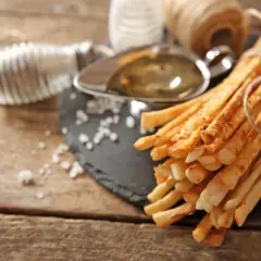 Grissini fatti in casa dorati, legati con spago, su un tagliere con sale grosso, olio e un rotolo di filo metallico.
