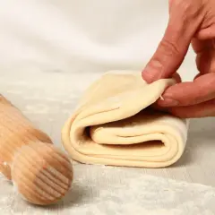 A hand folds layers of puff pastry dough next to a wooden rolling pin on a floured surface.