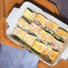 Involtini di zucchine pronti per essere cotti, ripieni e impanati, in una teglia bianca su un tovagliolo arancione.