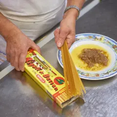 Mani che preparano i bigoli in salsa, con la pasta che esce dalla scatola e la salsa pronta nel piatto.