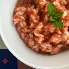 Un cremoso riso al pomodoro, guarnito con una fogliolina di menta fresca, servito in una ciotola bianca su una tovaglia colorata.