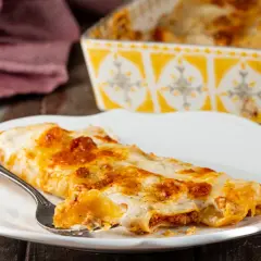 Piatto di cannelloni al forno fumanti, ripieni di carne e ricoperti di besciamella e sugo. Una forchettata mostra il ripieno invitante.