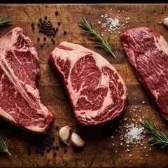 Tre succulente costate di manzo, pepe, sale, aglio e rosmarino su un tagliere di legno rustico, pronte per essere cucinate.