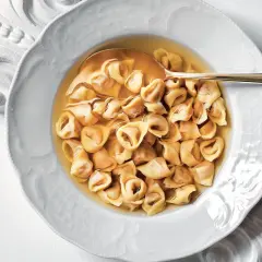 Un piatto di tortellini in brodo, un classico confortante, servito in una ciotola bianca decorata con un cucchiaio dorato.