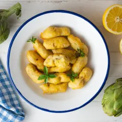 Carciofi fritti dorati, preparati con una pastella croccante, serviti in un piatto bianco con limone e carciofi freschi.