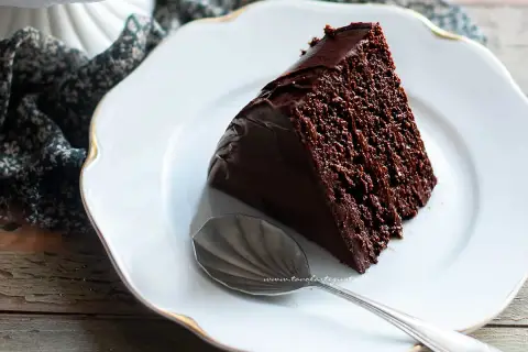 Una fetta di torta al cioccolato, umida e golosa, su un piatto bianco con bordo dorato. Accanto, un cucchiaino d'argento.