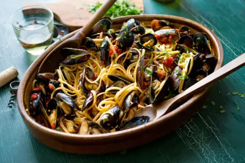 Un invitante piatto di pasta con le cozze, condita con pomodoro e prezzemolo, servita in una ciotola di legno con cucchiai.