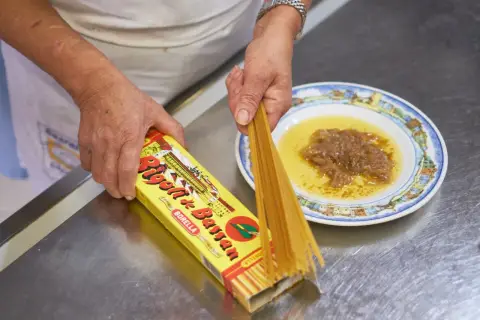 Mani che preparano i bigoli in salsa, con la pasta che esce dalla scatola e la salsa pronta nel piatto.