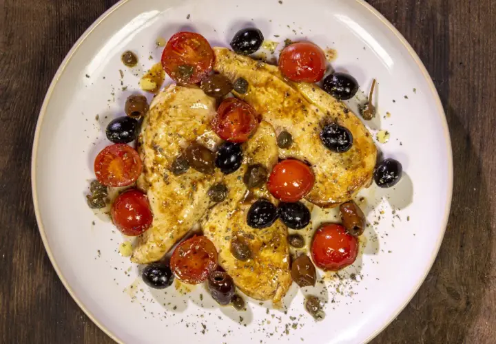 Petto di pollo in padella con pomodorini, olive nere e capperi, condito con erbe aromatiche.