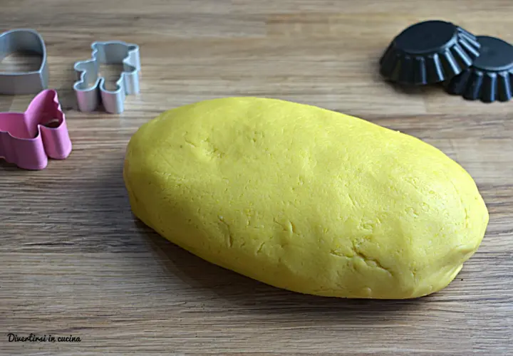 Yellow pasta frolla dough with cookie cutters and tart molds on a wooden surface.