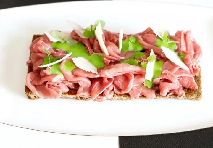 Delicato carpaccio di manzo su crostino integrale, guarnito con crema verde, scaglie di parmigiano e germogli freschi.