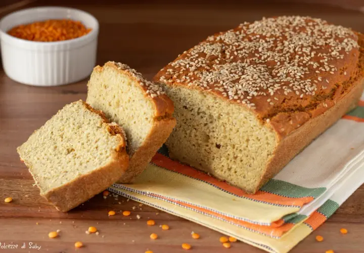 Pane di lenticchie dorato e soffice, cosparso di semi di sesamo, con due fette tagliate. Sullo sfondo, una ciotola di lenticchie rosse.