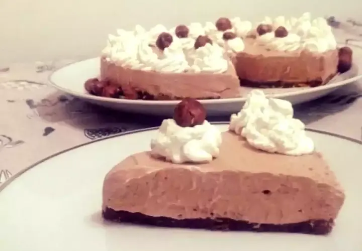 Una fetta di golosa cheesecake alla Nutella, guarnita con panna montata e nocciole intere, su un piatto bianco.