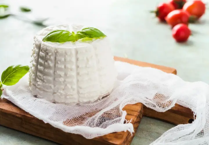 Ricotta fatta in casa, fresca e profumata, guarnita con basilico su un tagliere di legno. Pomodorini rossi sfocati sullo sfondo.
