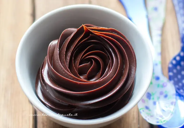 Una ciotola bianca piena di ganache al cioccolato, con un vortice perfetto. Vicino, cucchiai colorati.