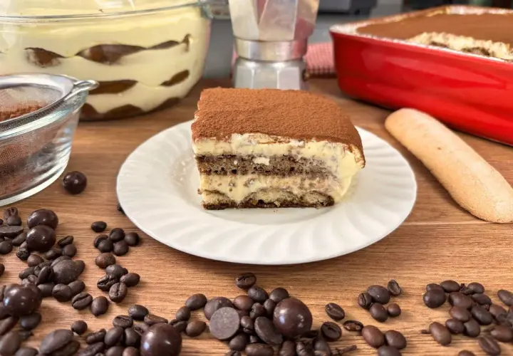 Una fetta di tiramisù con uova pastorizzate, cosparso di cacao, su un piatto bianco, con chicchi di caffè e cioccolatini sparsi.