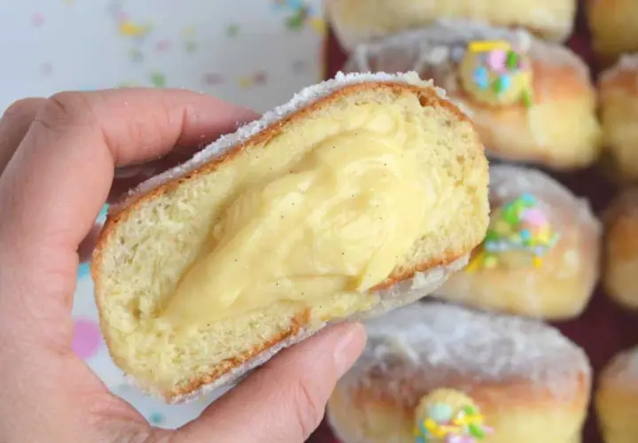 Un bombolone alla crema, soffice e ripieno, viene tenuto in mano. Altri bomboloni alla crema decorati con confettini colorati sono sullo sfondo.