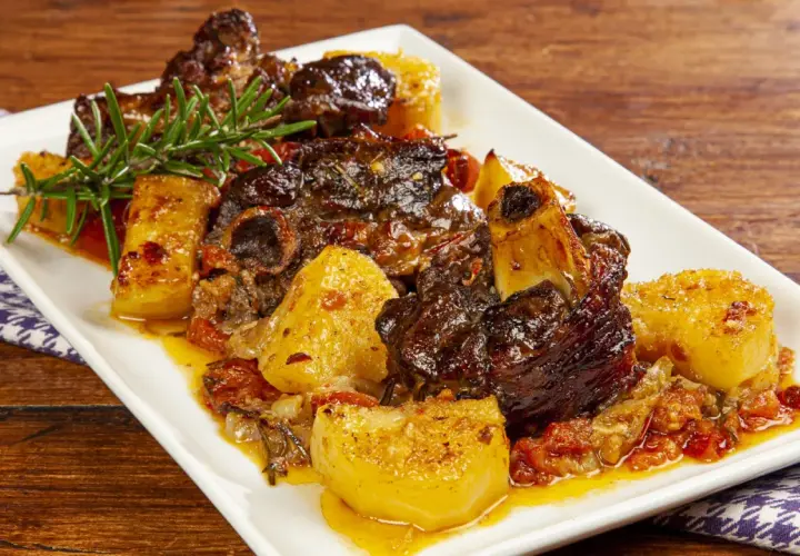 Un succulento agnello al forno con patate dorate, pomodorini e rosmarino su un piatto bianco.