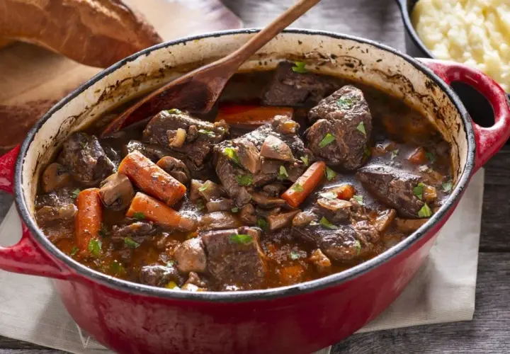 Un invitante spezzatino di manzo in un tegame rosso, con carote, funghi e pane rustico sullo sfondo.