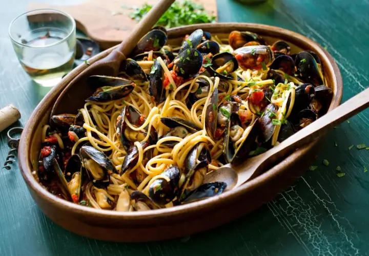 Un invitante piatto di pasta con le cozze, condita con pomodoro e prezzemolo, servita in una ciotola di legno con cucchiai.