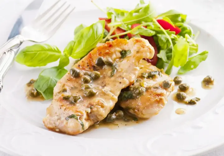 Deliziose scaloppine di pollo al limone e capperi, servite con un'insalata fresca e foglie di basilico.