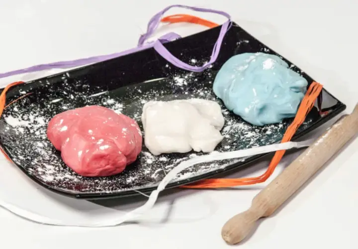 Tre panetti di pasta di zucchero colorata (rosa, bianca, azzurra) su un piatto nero con nastri colorati e un mattarello.
