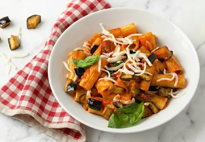 Un piatto di pasta con melanzane, pomodoro e formaggio grattugiato, guarnito con basilico fresco. Accanto, un tovagliolo a quadri rossi e bianchi.