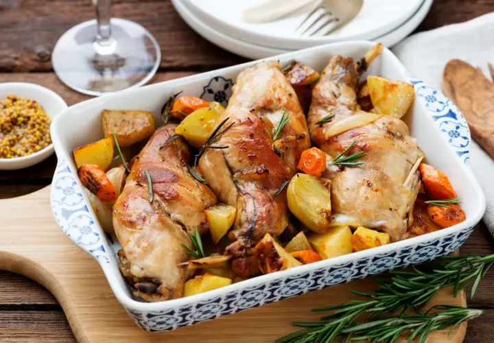 Coniglio al forno dorato con patate e carote, profumato al rosmarino, servito in una pirofila decorata.