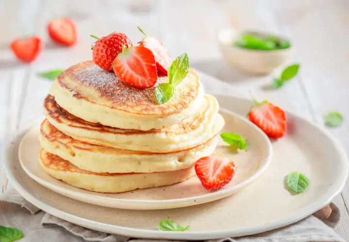 Pancake allo yogurt soffici e dorati, guarniti con fragole fresche e zucchero a velo. Un dolce inizio di giornata.