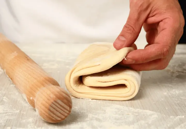 A hand folds layers of puff pastry dough next to a wooden rolling pin on a floured surface.