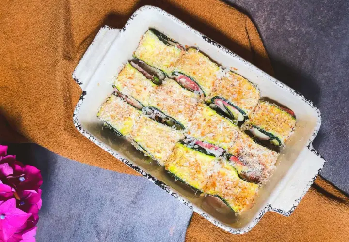 Involtini di zucchine pronti per essere cotti, ripieni e impanati, in una teglia bianca su un tovagliolo arancione.
