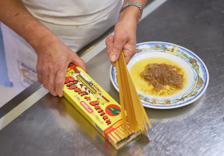 Mani che preparano i bigoli in salsa, con la pasta che esce dalla scatola e la salsa pronta nel piatto.