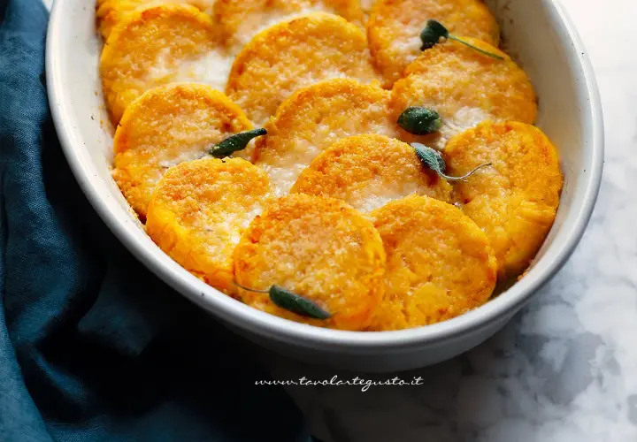 Gnocchi di zucca dorati e gratinati, guarniti con foglie di salvia, in una pirofila bianca.