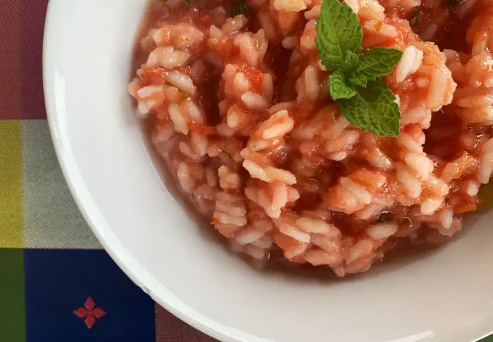 Un cremoso riso al pomodoro, guarnito con una fogliolina di menta fresca, servito in una ciotola bianca su una tovaglia colorata.