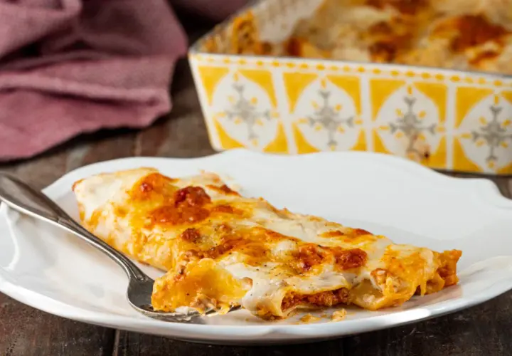 Piatto di cannelloni al forno fumanti, ripieni di carne e ricoperti di besciamella e sugo. Una forchettata mostra il ripieno invitante.