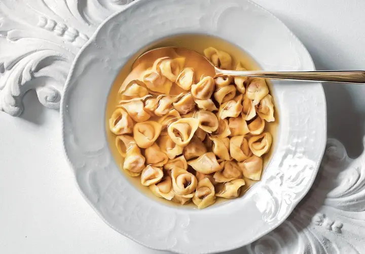Un piatto di tortellini in brodo, un classico confortante, servito in una ciotola bianca decorata con un cucchiaio dorato.